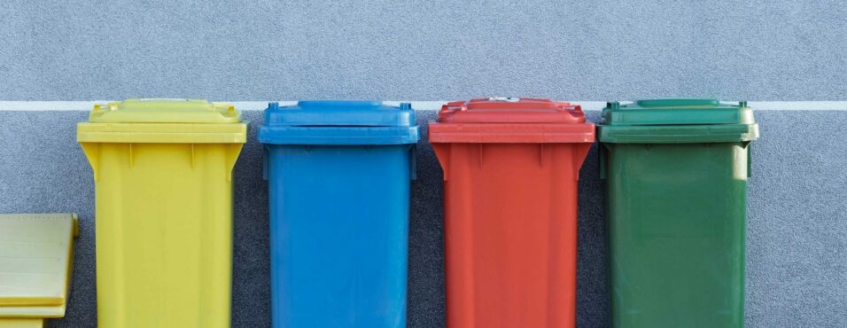 four assorted-color trash bins beside gray wall
